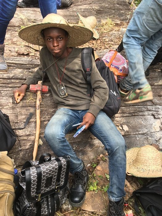 A student in a straw hat seated outdoors with a hammer and field bag